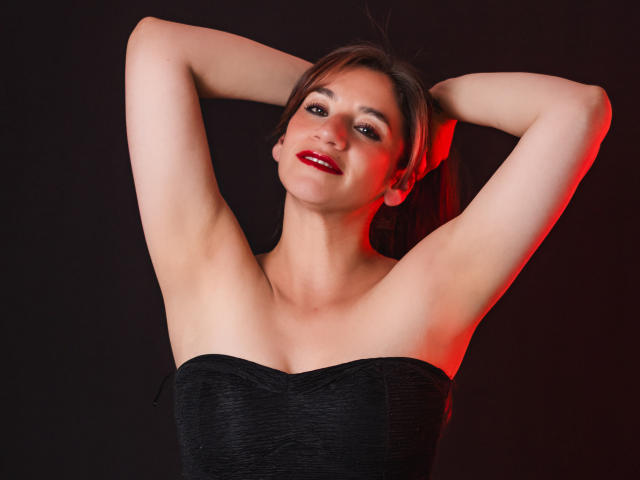 AvaStone con le braccia alzate, che mostra le ascelle scoperte, indossa un abito nero senza spalline, rossetto rosso e un leggero sorriso, su uno sfondo scuro con illuminazione rossa..