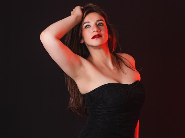 AvaStone, con cabello castaño largo, lápiz labial rojo y un vestido negro sin tirantes, levantando su brazo derecho, mostrando su axila y sus pequeños senos firmes, sobre un fondo oscuro..