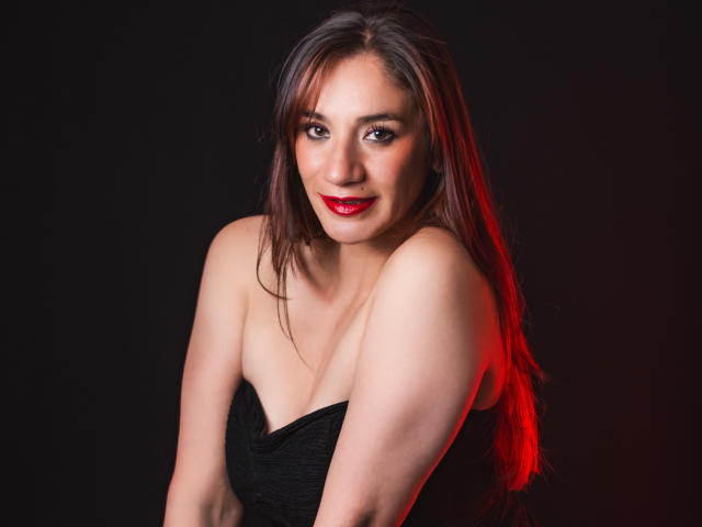 AvaStone con lápiz labial rojo, vestido negro sin tirantes y cabello castaño largo, sonriendo a la cámara, mostrando sus senos y hombros desnudos sobre un fondo oscuro..