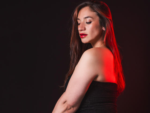 AvaStone, con le labbra rosse, gli occhi chiusi e un'espressione sensuale, indossa un abito nero senza spalline, con un'illuminazione rossa laterale e ha i capelli lunghi e castani, mostrando la spalla e il braccio scoperti. Il suo seno è leggermente visibile..