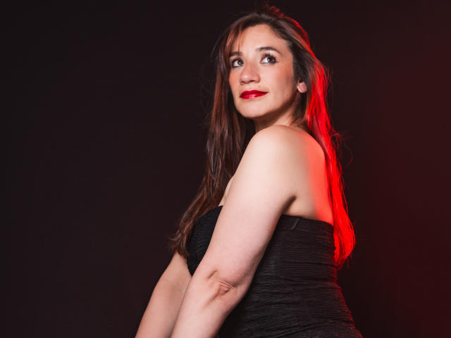 AvaStone con lápiz labial rojo, vestido negro sin tirantes y cabello castaño largo, mirando por encima del hombro, luz roja a la derecha, brazos desnudos, ligera sonrisa, fondo oscuro..