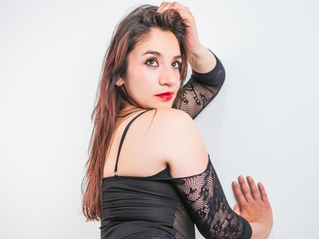 AvaStone with long brown hair, red lipstick, and black lace off-shoulder top, looking over her shoulder, hand in her hair, showing her bare back and black bra strap.