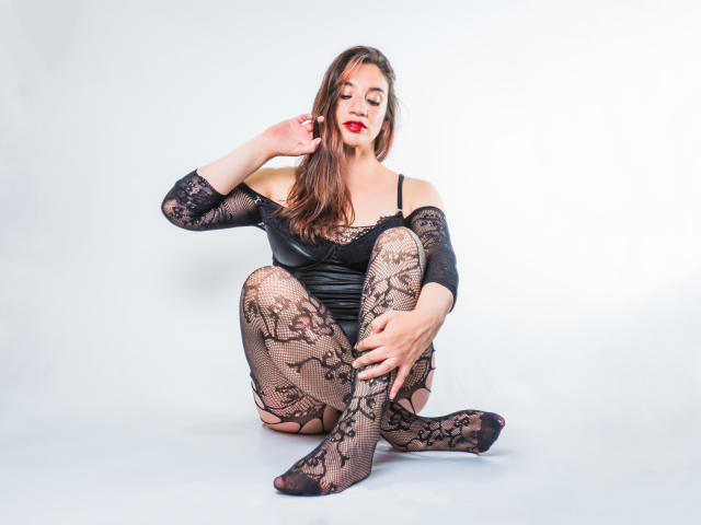 AvaStone with long brown hair, red lipstick, black lace lingerie, and fishnet stockings, sitting on the floor, hand near ear, looking down, legs crossed, showing her tits.