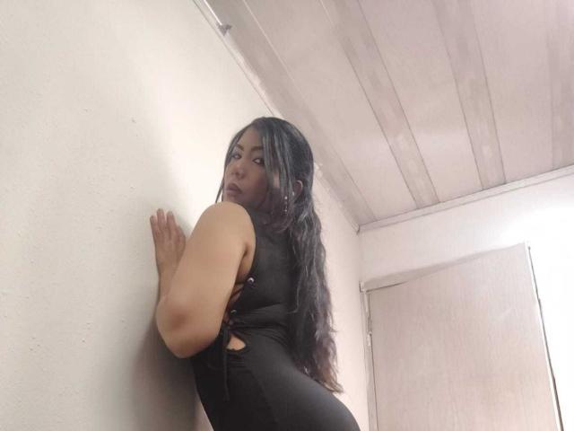 IsadoraVex with long black hair, wearing a tight black dress, leaning against a white wall, looking back with a slightly open mouth, showing her ass.