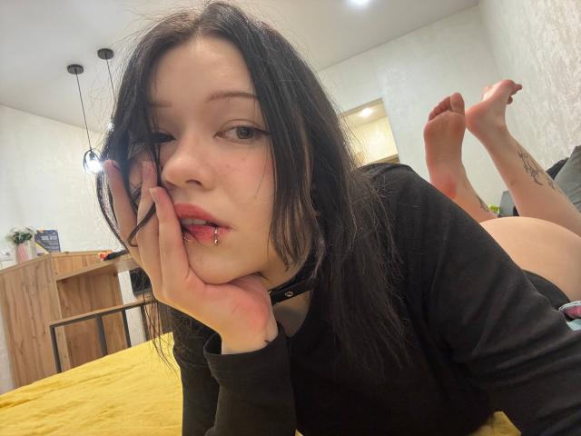 Delora Detzler mit schwarzen Haaren, die ein schwarzes Hemd trägt und auf einem gelben Bett liegt, beißt sich in den Finger, hat ein Lippenpiercing, die Beine sind hochgehoben, und ihr nackter Hintern ist sichtbar..