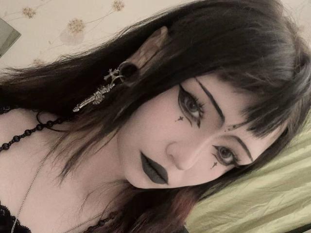 Deloras Detzler, met donkere make-up, zwarte lippenstift, goth eyeliner, een ketting van zwarte kralen, zwart haar, oorbel piercings en een oorbellen in de vorm van een schedel, kijkt naar de camera met bleke huid..