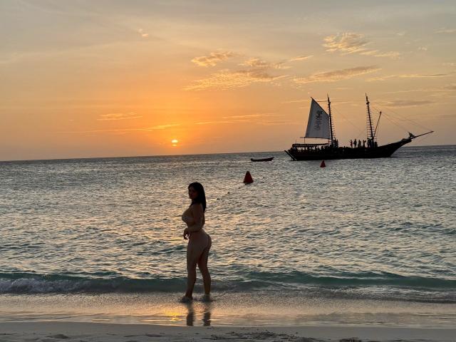 MaddieDemie stands in the shallow water, nude, with her ass and tits visible, watching a sunset and a black ship with sails on the horizon.