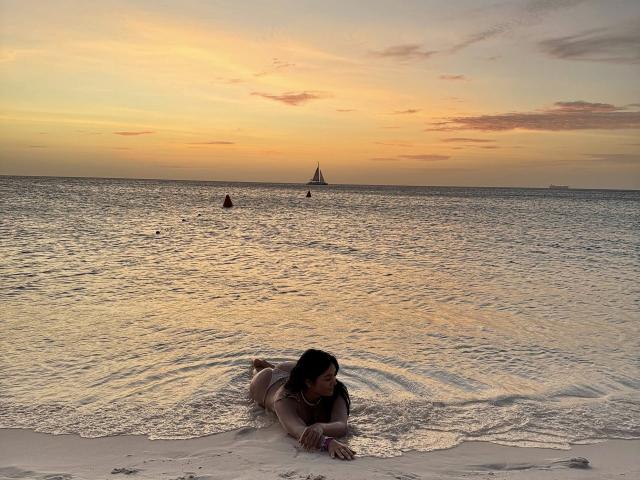 MaddieDemie deitada em uma praia, de bruços, de biquíni, com o bumbum levantado e os seios levemente visíveis, olhando para a água, ao pôr do sol..