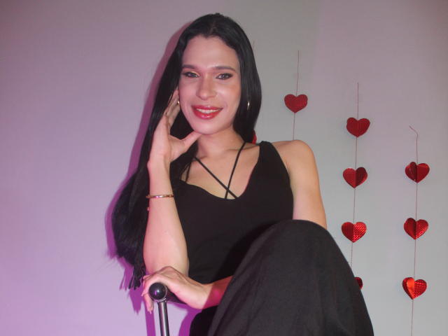 JadeOconner with long black hair, red lipstick, black sleeveless top, and black pants, sitting on a chair, smiling, hand on cheek, red heart decorations on wall.