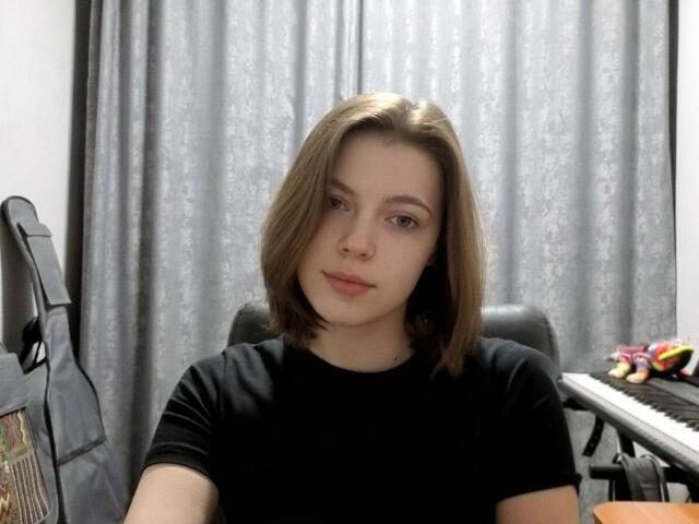JennellMatacale, a young woman with shoulder-length brown hair, wearing a black shirt, sitting in front of a keyboard and a guitar, with a neutral expression.