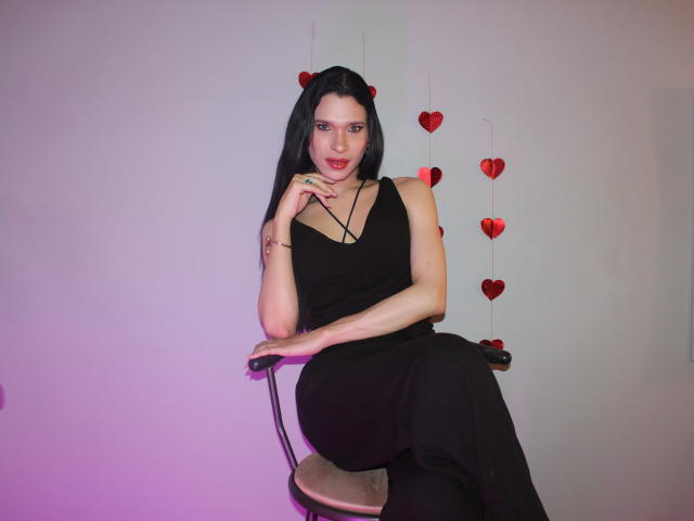 JadeOconner with long black hair, wearing a black sleeveless dress, sitting on a chair, hand near mouth, red heart decorations on wall, pink-purple light.