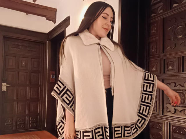 MabyLegrand with long brown hair, wearing a white cape with black Greek pattern, pink top, black pants, smiling softly, standing near a dark wooden door and patterned cabinet.
