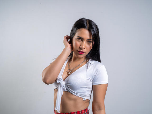 VelvetRoss with long black hair, wearing a tied white crop top showing her small tits and a red plaid skirt, holding her ear with a serious expression.
