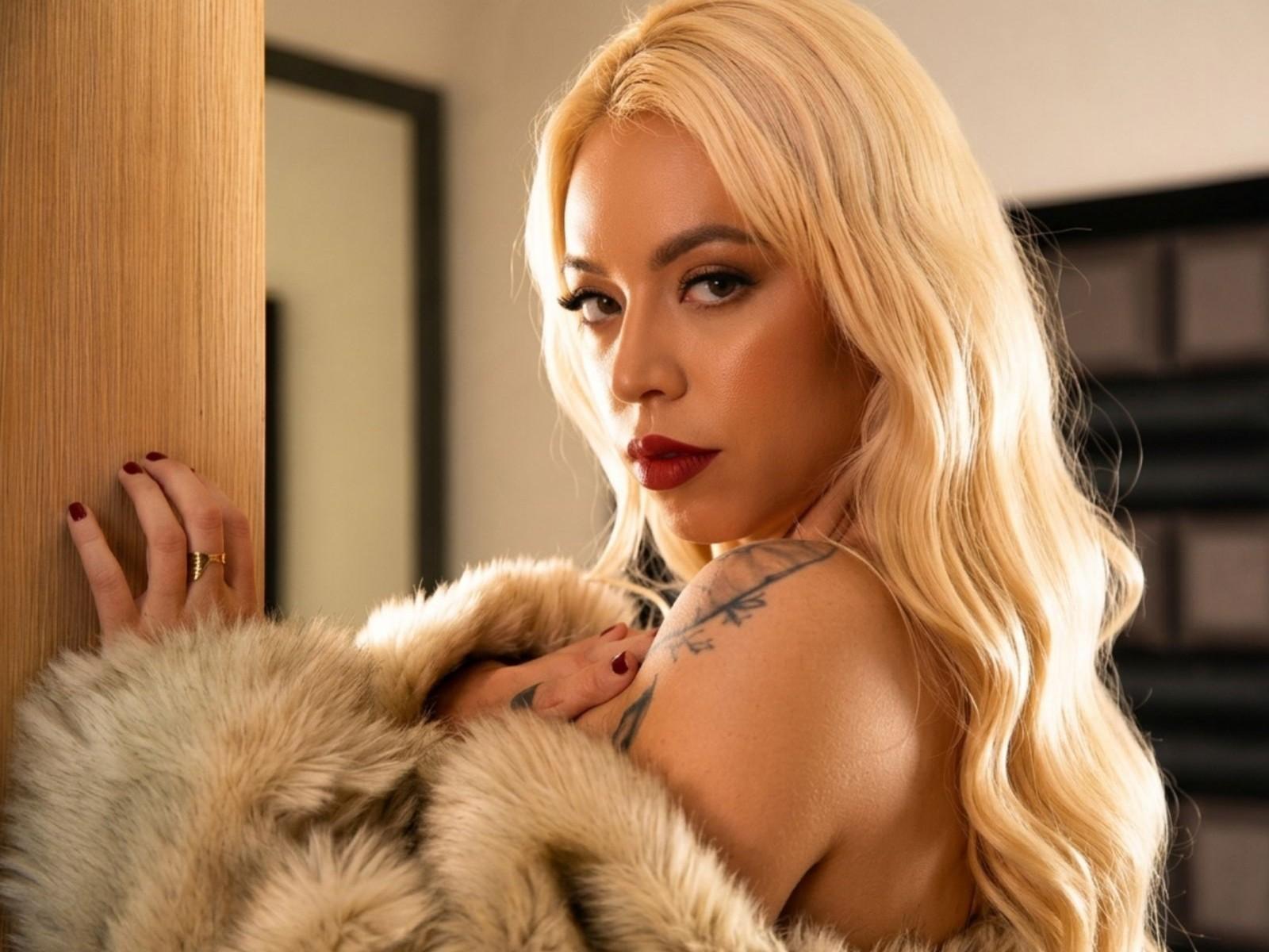 Natashha: Blond haar, rode lippenstift en tatoeages, draagt een bontjas, kijkt verleidelijk, raakt een houten muur aan, nagels zijn donkerrood gelakt, gouden ring om de vinger..