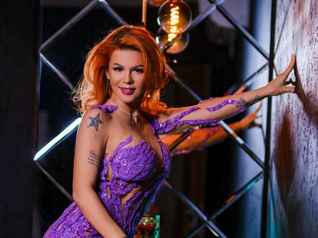 CamillaStarX, met oranje haar, draagt een paarse, pailletten jurk, waarbij haar borsten zichtbaar zijn. Ze heeft sterren-tatoeages op haar schouder en leunt tegen een muur met een verleidelijke glimlach..