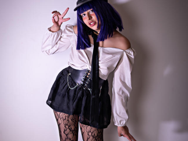 Carly Blake, avec sa coupe de cheveux courte bleue, son chemisier blanc à épaules dénudées, sa jupe noire, ses bas en dentelle et sa ceinture à chaîne, levant le majeur, portant un chapeau noir, exhibant ses seins et ses fesses..
