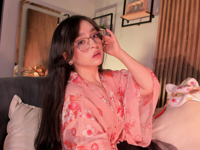 Eva McAllister, con lunghi capelli neri, indossa un kimono fiorato rosa, occhiali e orecchini, mentre si sistema gli occhiali, seduta su un divano con un cuscino e una coperta..