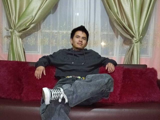 AdonisLatin with short black hair, dark hoodie, loose jeans, and white sneakers, sitting on a red couch, legs crossed, arms resting, green curtains behind, slight smile.