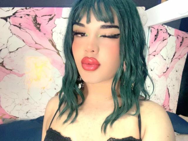 GlossyyBratz with teal hair, black lace top, and red lips, winking with long eyelashes, has pouty ass-kissing lips, and a cute, sexy, and attractive look.