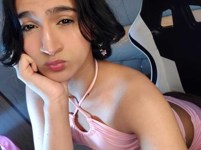 ZoeKiiss with dark wavy hair, pink halter top, and pink shorts, pouting with nose ring, hand on cheek, looking at camera, showing small tits and cute pink lips.