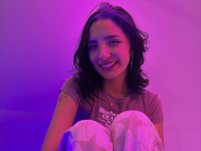 ArisSuarez with dark wavy hair, smiling, wearing a gray shirt and white patterned pants, sitting with knees up, purple and pink lighting, visible tattoo on right arm.