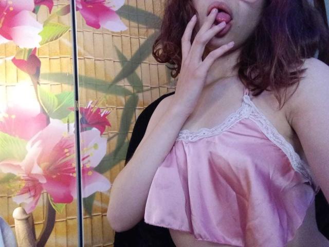 Sophie Blake, con i capelli rossi ondulati, indossa una camicetta rosa in raso con pizzo, ha la lingua fuori, le dita che toccano la bocca, mostrando il seno, in una camera da letto con decorazioni floreali alle pareti..