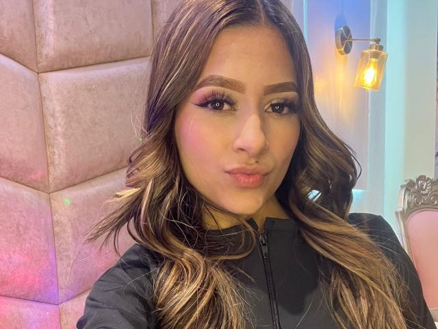 AfroditaWild with wavy brown hair, pink eyeshadow, and pouty lips, wearing a black zip-up shirt, has a cute, sexy face, and a small, pretty nose.