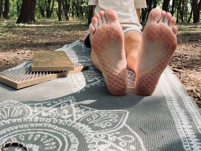 TenerliRize's bare soles, pinkish with tiny bumps, facing camera, feet up, on a grey patterned mat, wooden comb to left, forest background, daytime, TenerliRize in white shirt.
