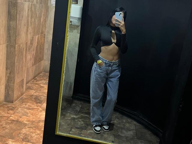SaraMillerOne, con il seno scoperto, indossa una canottiera nera, jeans blu e scarpe nere, e sta scattando un selfie allo specchio in un bagno. La sua mano è infilata nella tasca dei jeans..