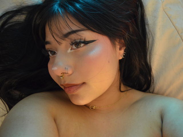 Annie Russel mit dunklem Haar, Eyeliner, Nasenring, goldener Halskette und Ohrringen, die in die Kamera lächelt, oben ohne, wobei ihre Brüste zu sehen sind. Weiches Licht auf ihrem Gesicht..