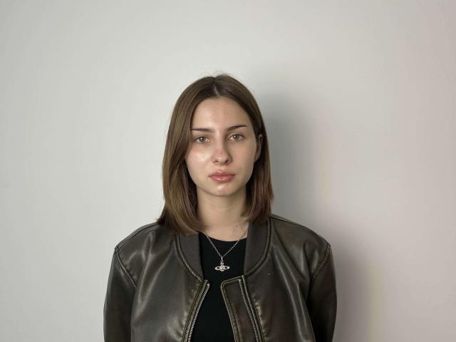 NellieHeimer with medium brown hair, green eyes, and a serious expression, wearing a black leather jacket and a silver necklace with a small pendant, against a plain white background.