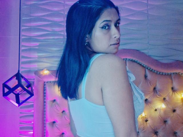 RoxanneHan met lang donker haar, een witte tanktop, kijkt om, waarbij haar achterkant zichtbaar is. Ze zit op een gestoffeerde, roze-gele, met studs versierde hoofdbord met paarse en roze verlichting..