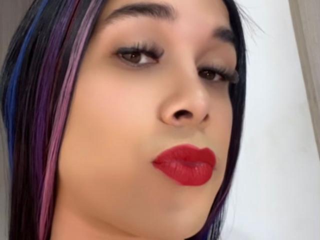 O rosto de VanessaCandyHot, com batom vermelho, sobrancelhas escuras, olhos castanhos e cabelo preto com mechas roxas. Ela está olhando ligeiramente para o lado, com um leve sorriso..