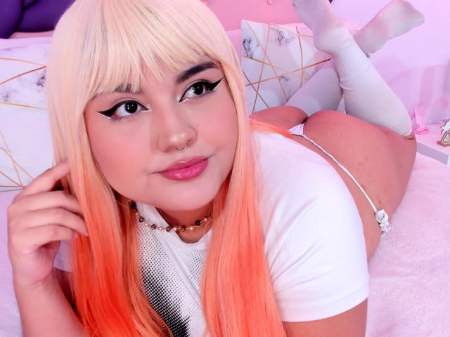 MarlaBriard with blonde-orange hair, white eyeliner, nose ring, white shirt, white panties, white socks, lying on bed, showing her ass, smiling, pink lipstick, soft pink lighting.