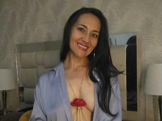 MiaMott, con lunghi capelli neri, sorridente, indossa una camicia blu aperta che lascia intravedere il seno, ornato da una collana con un fiore rosso, seduta in una camera da letto scarsamente illuminata..