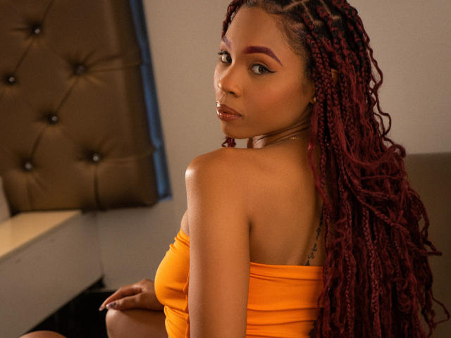 SkinnySamantha, com cabelo trançado vermelho escuro, vestindo um top laranja sem alças, olhando por cima do ombro, mostrando suas costas nuas e uma tatuagem no braço superior..