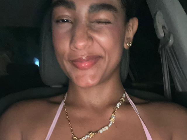 NatalyBlonder with one eye closed, smiling, pink halter top, gold and bead necklace, small gold earrings, dark hair, dimly lit, car interior, slight drool on her chin.