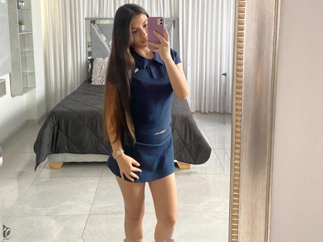 IsabelaRios in a navy blue, short-sleeve, athletic top and matching mini-skirt, standing in front of a mirror, holding a pink phone, with long brown hair, showing her ass.