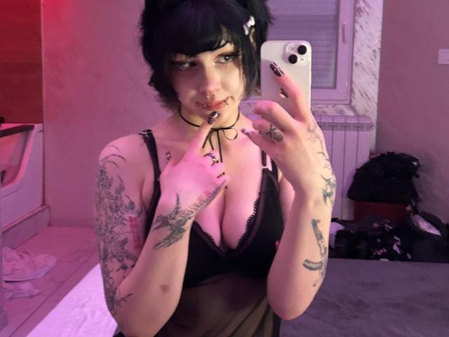 AshleyAlt with black hair, tattoos, and piercings, holding a phone, wearing a black lace bra, showing her boobs, and a choker, in a pink-lit room.