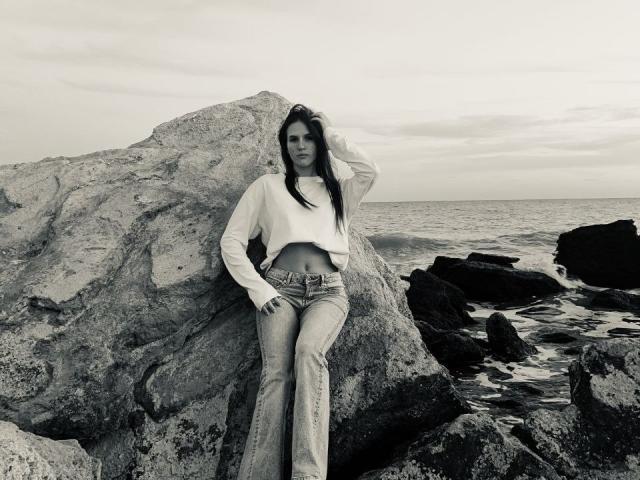 OliviaaShinee, avec de longs cheveux noirs, un débardeur blanc et un jean clair, appuyée contre un grand rocher, une main dans les cheveux, l'océan et les rochers en arrière-plan, photo en noir et blanc..