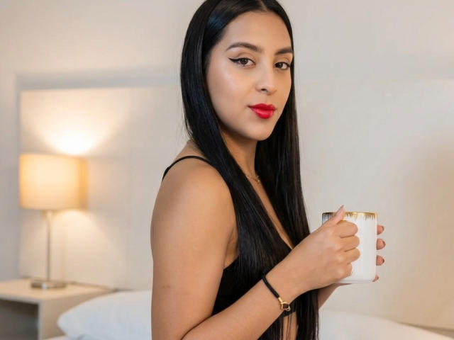 EmmaEstreilla, met lang zwart haar, rode lippenstift, die een witte mok vasthoudt, gekleed in een zwarte jurk, staat in een slaapkamer en kijkt met een lichte glimlach naar de camera..