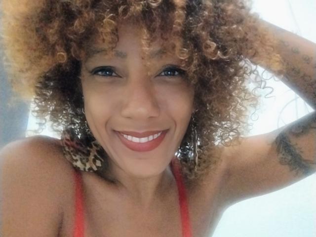 CinnamonBebe has curly blonde hair, blue eyes, and a big smile. She's wearing red lingerie with her hand in her hair, showing a tattoo on her arm.