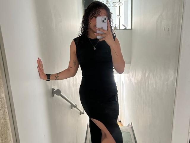 SofiaPassionne, con cabello rizado, vestida con un vestido negro, mostrando un corte en la pierna, sosteniendo un teléfono, tatuaje en el brazo, selfie frente al espejo, pasillo blanco, pulsera negra..