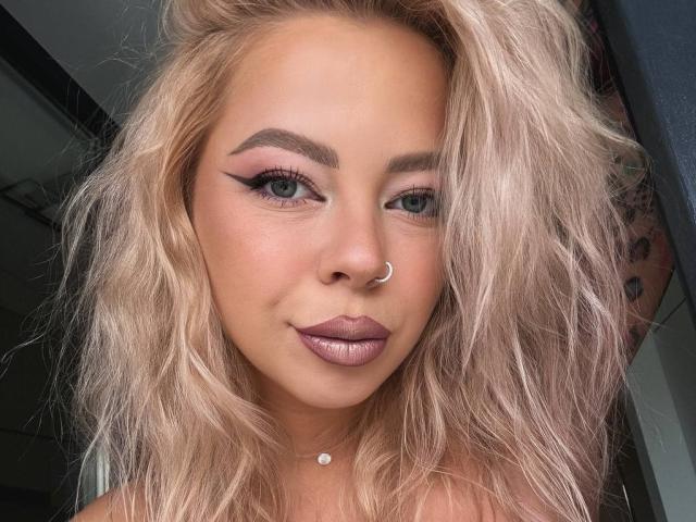 A RedLara tem cabelo loiro ondulado, olhos verdes, delineador marcante, batom roxo e um piercing no nariz. Ela está sorrindo e usa um colar pequeno com um diamante. Seus seios não estão visíveis..