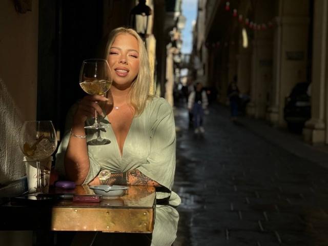 RedLara with blonde hair, green dress, holding a glass of white wine, smiling, sitting at a table with two empty wine glasses, sunlight on her, cobblestone street.