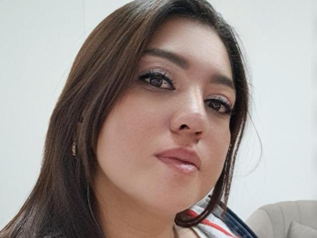 PaulaAnd tem cabelo castanho, delineador escuro e lábios rosa brilhantes. Seus olhos castanhos olham ligeiramente para o lado com um sorriso discreto. Ela está vestindo uma camisa listrada..