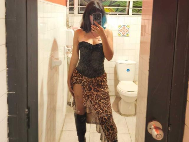 KleoLennox with black lace strapless top, leopard print skirt with high slit, and black knee-high boots, taking a mirror selfie in a bathroom, showing her tits.