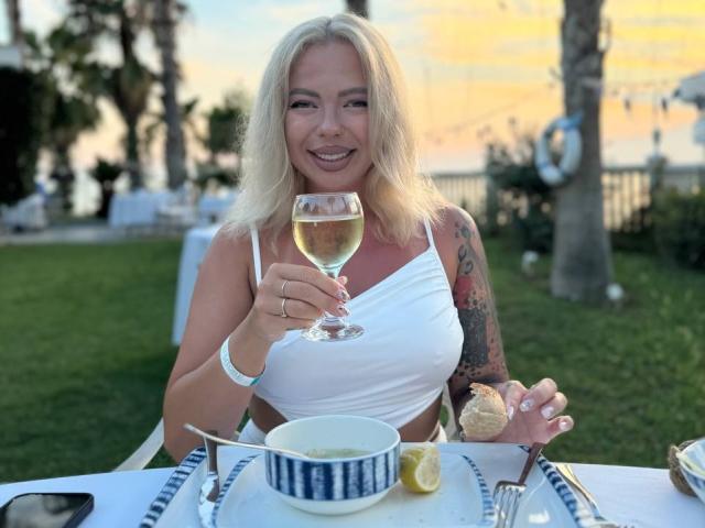 RedLara with blonde hair, white tank top, and tattoos, holding a glass of white wine, smiling, sitting outdoors, table with food, lemon, and cutlery in front.