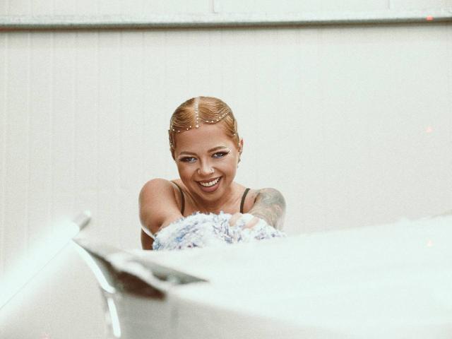 RedLara with blonde hair, a head chain, and tattooed arm, smiling and holding a fluffy white bouquet, leaning over a white surface, looking up at the camera.