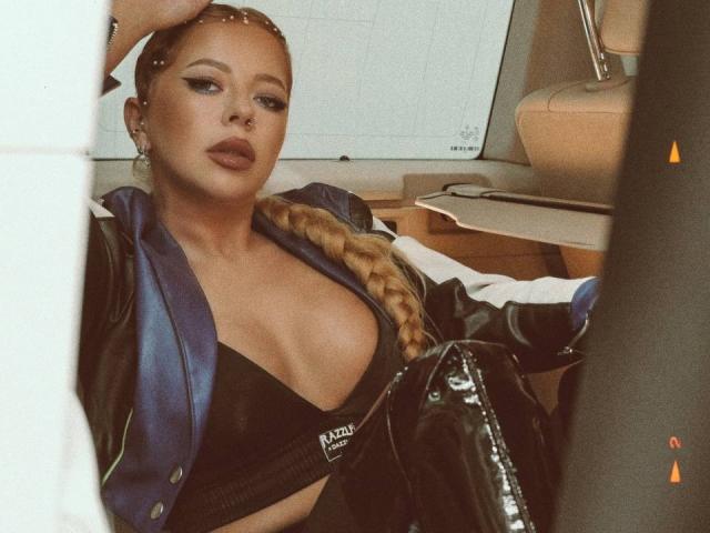 RedLara, avec des tresses blondes, portant une veste en cuir bleu et un haut noir, laissant entrevoir ses seins volumineux, assise dans une voiture, regardant l'appareil photo avec une expression confiante..